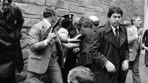 Secret Service agents and police officers swarm John Hinckley, obscured from view, after he attempted an assassination on President Ronald Reagan.