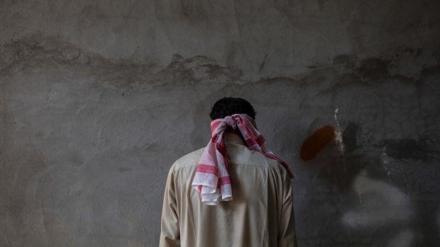 A blindfolded Islamic State group suspect stands against a wall facing away from Kurdish security officers.