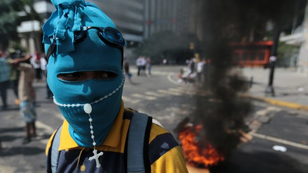 A youth wearing a mask and rosary stands near a burning barricade  in Caracas.
