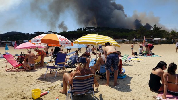 People enjoy sunbathing on the beach in Lavandou, French Riviera, as plumes of smoke rise in the air from burning wildfires.