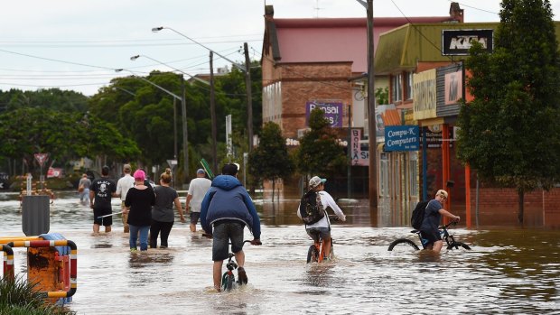 Teenagers cycle behind a group of people with brooms wading through the water of the flooded streets of Lismore.