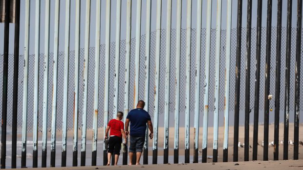 Two people walk towards metal bars marking the United States border where it meets the Pacific Ocean in Tijuana, Mexico.