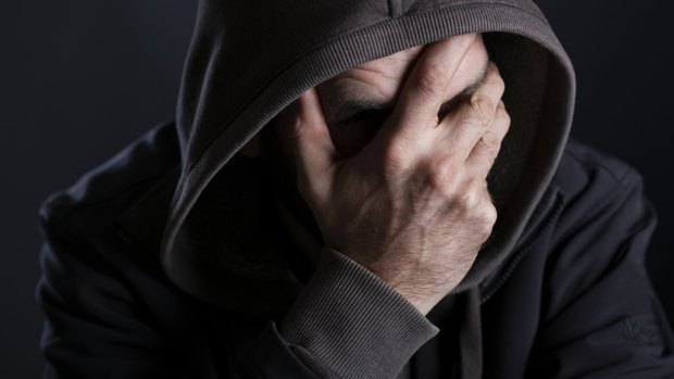 Depressed man looking despaired, hiding face with hand and hood, isolated on black background.