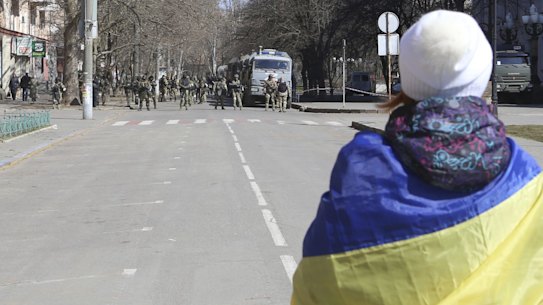 A woman covered by Ukrainian flag stands in front of Russian troops in a street during a rally against Russian occupation in Kherson, Ukraine, Saturday, March 19, 2022. (AP Photo/Olexandr Chornyi)