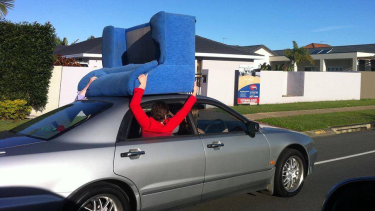 Two passengers hold a couch on the roof of a moving car.