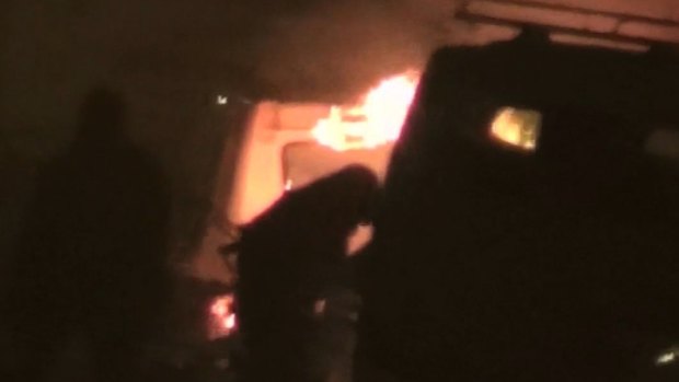 Two unidentified armed men approach a burning vehicle near a hotel in Ouagadougou, Burkina Faso.
