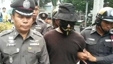 A man in a Guy Fawkes mask and sunglasses is escorted by Thai police in Bangkok. 