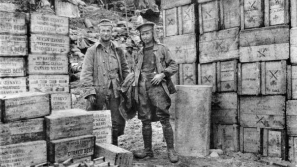 Two unidentified soldiers standing on the beach at Anzac Cove on the Gallipoli Peninsula in 1915, flanked by boxes of bully beef.