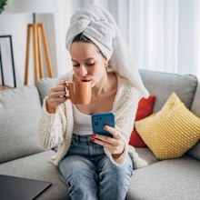 One woman, beautiful young woman sitting on sofa in living room and using smart phone, she has a towel on her head.