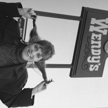 Wendy Morse, daughter of Wendy's fast-food chain founder Dave Thomas, holding up her hair in simulated pigtails under a Wendy's sign