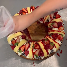 Woman adding home-made whipped cream between the alternating vanilla scrolls with jam and lamingtons sitting on a wooden plate in preparation for a Christmas "wreath" dessert.