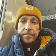 jonnie irwin sitting on a plane smiling and wearing a beanie