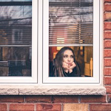 Young woman in lockdown, sitting inside her home, close to the window looking out. She is practicing social distancing due to COVID-19