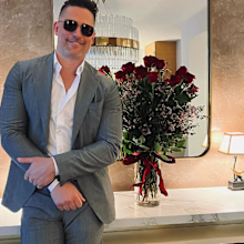 Joe Manganiello posing in a suit with a bunch of red roses in a vase.