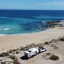 A car with attached caravan is parked on the sand next to a stunning blue beach. Auswide Explorers Instagram