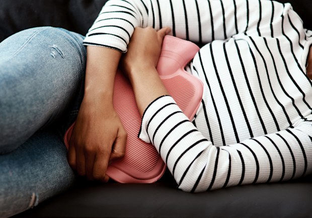 Cropped shot of a woman holding a hot water bottle against her stomach on the sofa at home