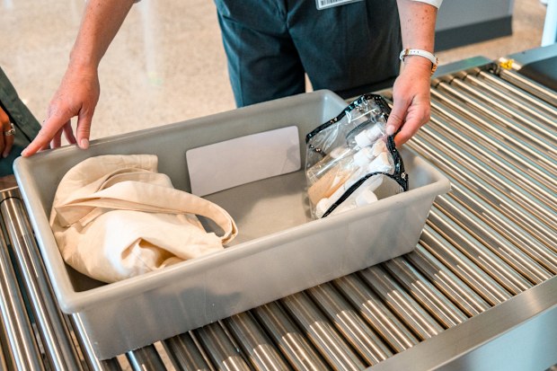 Female security employee holding a plastic tray with  female traveler's personal items, before pushing it through the baggage scanner. Only arms of women visible.