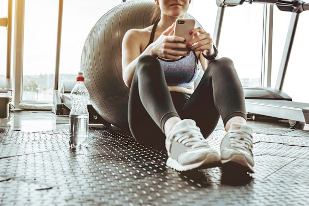 Young woman athlete using cell phone at gym. Young woman in sportswear checking phone while resting after workout on floor. Beautiful fit girl messaging with smartphone at fitness centre.