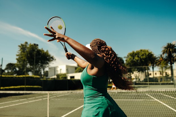 Female tennis player seen from behind, hitting a volley on a hardcourt during a sunny day. Sporty woman playing tennis outdoors.