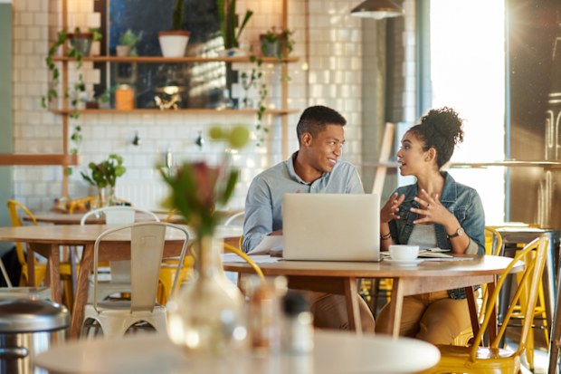Cropped shot of two young business colleagues using a laptop during a discussion over their financial paperwork in a cafe