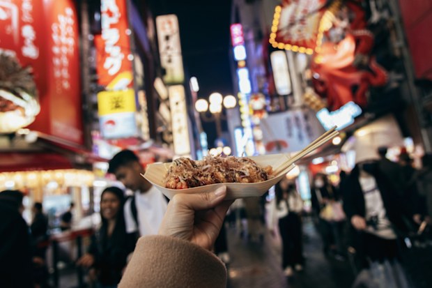 The bustling city streets serve as the backdrop, filled with vibrant shops, colorful signage, and a multitude of people moving about. The sounds of chatter and street performers create an energetic atmosphere.