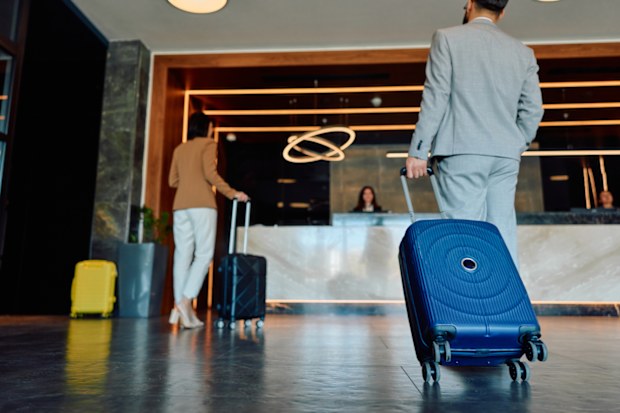 Business travelers arrive at a modern hotel lobby, pulling suitcases across the floor toward the reception desk as they prepare to check in and begin their stay
