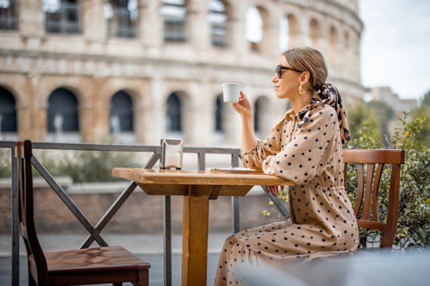 Woman drinking coffee at outdoor cafe near coliseum, the most famous landmark in Rome. Concept of italian lifestyle and traveling Italy. Caucasian woman wearing dress and shawl in hair