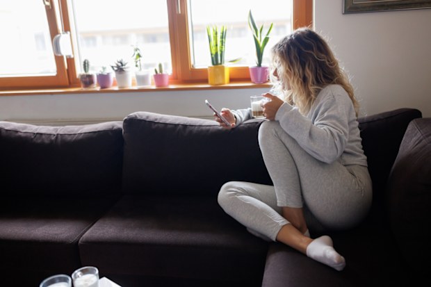 woman sitting on sofa, drinking coffee and texting on mobile phone at home