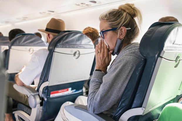 Woman sitting on a plane blowing her nose. Her protective face mask to reduce the spread of COVID-19 is under her chin. She is suffering from cold and flu symptoms.