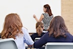 Rear view of primary school teacher writing on a clear whiteboard in classroom with students.