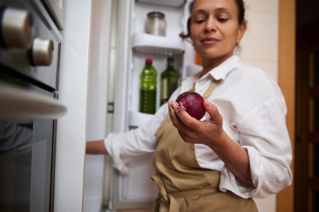 A woman in a kitchen apron holds a red onion near an open refrigerator, preparing ingredients for cooking.