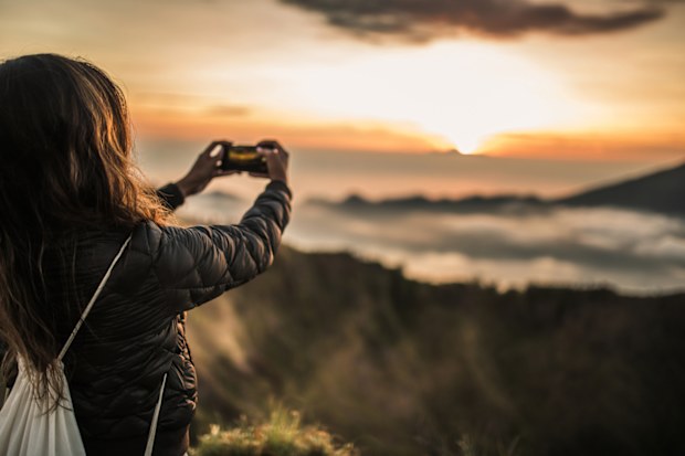 One woman, standing high on beautiful mountain Batur in sunset alone, photographing sunset with smart phone.