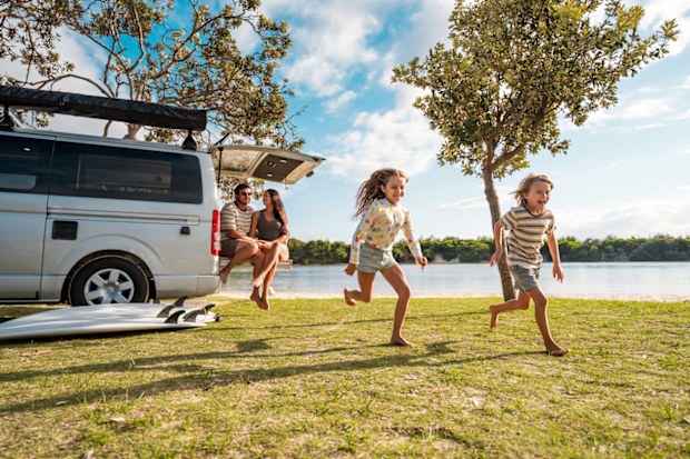 Wide-angle shot of young parents and children running playfully on grass by seaside campsite. White camper van parked under trees creates perfect outdoor recreation scene in natural summer setting.