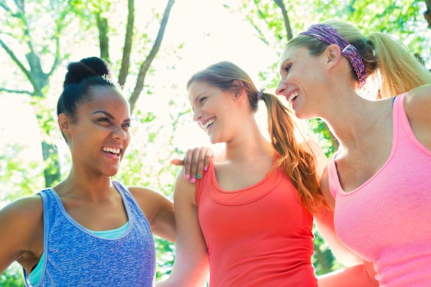 Young adult African American woman is smiling while talking to her mid adult Caucasian female athletic friends. Women are exercising together in sunny park with green trees. They are wearing sports tank tops in bright colors.