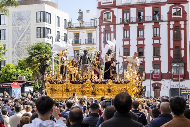 Easter Semana Santa procession in Seville, Spain, April 15, 2025. A golden religious float with biblical figures parades through crowded streets, surrounded by spectators and historic Andalusian buildings.