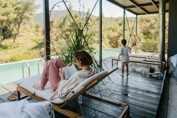 Photo of two boys having fun on a summer vacation in a house with a swimming pool, while their mom is relaxing over the book she is reading in a sun bed