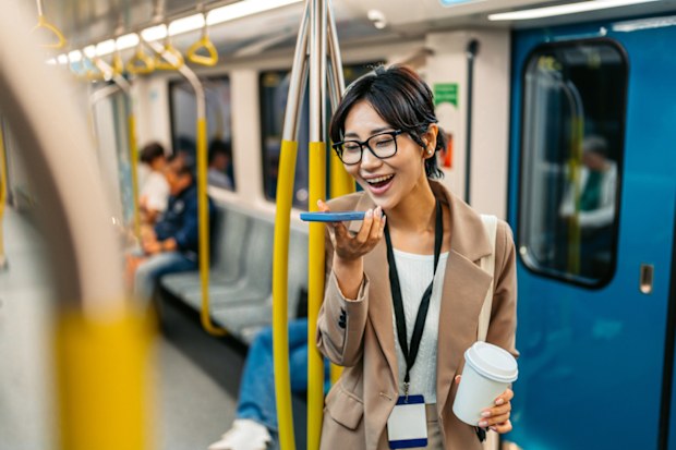 Beautiful young Japanese Asian businesswoman sending a voice message using her phone and drinking coffee to go while riding in a subway train in Sydney in Australia.