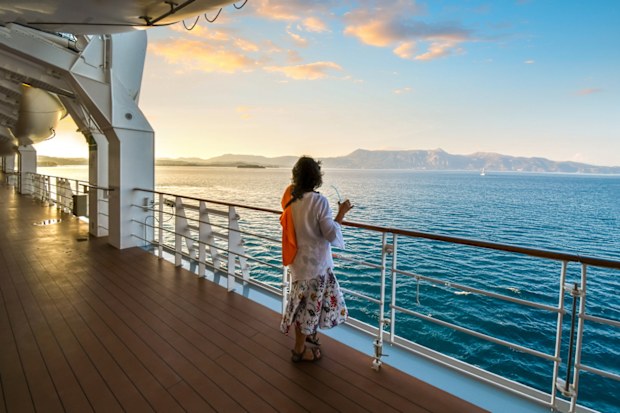 A woman sips a drink on the deck of a cruise ship as the sun sets and the ship passes islands on the Aegean Sea