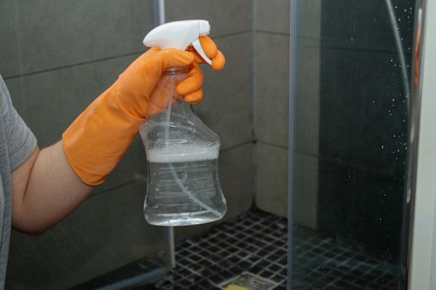 Close up of woman's hand in orange gloves cleaning dirty glass shower doors with lime remover spray
