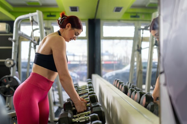 A woman in athletic wear is seen organizing dumbbells in a gym. The environment is equipped with modern fitness equipment creating a focused and vibrant atmosphere. The image captures a moment of preparation for strength training, symbolizing dedication, discipline, and fitness goals.