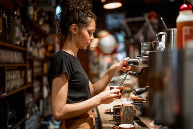 Aboriginal waitress in black attire operates professional espresso machine, demonstrating skilled coffee preparation techniques in a warmly lit cafe environment with traditional bar setup.