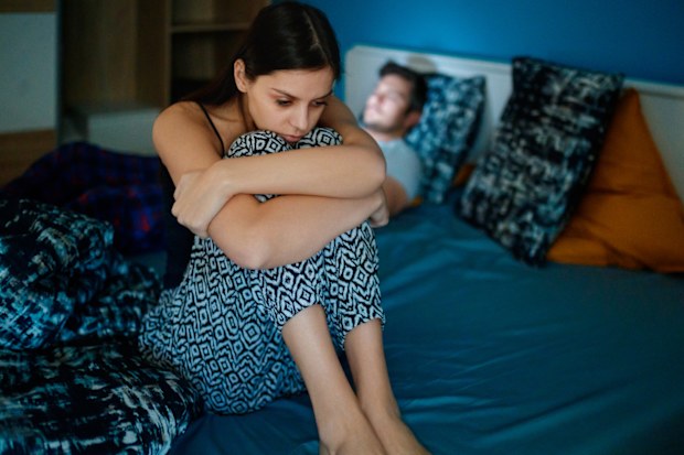 Sad girlfriend sitting on bed edge, holding her legs after fight, her partner is lying in bed in background, his face is lit by smart phone