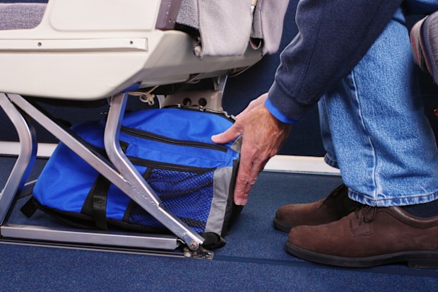 Airplane passenger stowing his carry-on luggage under the seat in front of him.