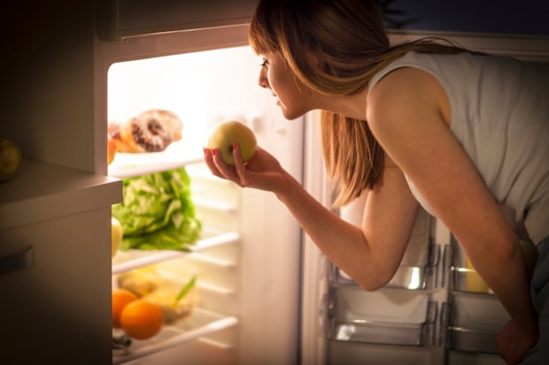 Close up image of a young woman, having a midnight snack - eating an apple, in front of the refrigerator.