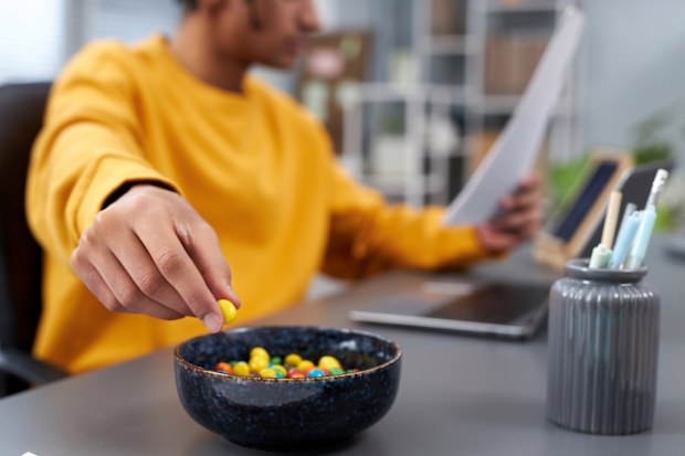 Close up of young man eating snacks while working or studying at home, focus on hand holding colorful candy
