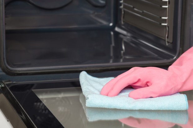 Woman hand in pink rubber protective glove using blue dry rag and wiping glass door surface inside oven at home kitchen.