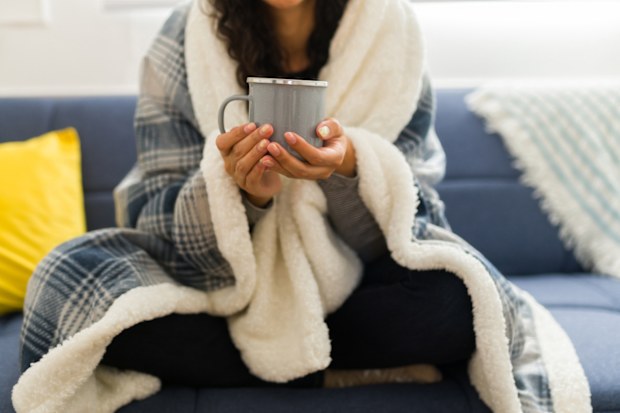 Close up of a young woman wrapped in a quilt relaxing on the sofa during autumn weather at home drinking hot tea or chocolate