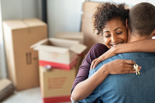 Young woman holding home keys while hugging boyfriend in their new apartment after buying real estate. Lovely girl holding keys from new home and embracing man. Happy couple in their apartment around cardboard boxes.