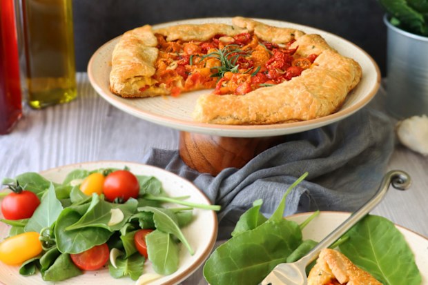 Stock photo showing close-up view of freshly cooked savoury galette, homemade, golden pastry crust, containing pesto, cherry tomato halves, crumbled feta cheese, sprinkled with rosemary leaves. Free form, on a white plate.