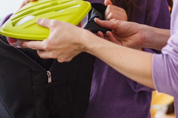 Mother and daughter packing lunch box into school backpack together at the kitchen counter.